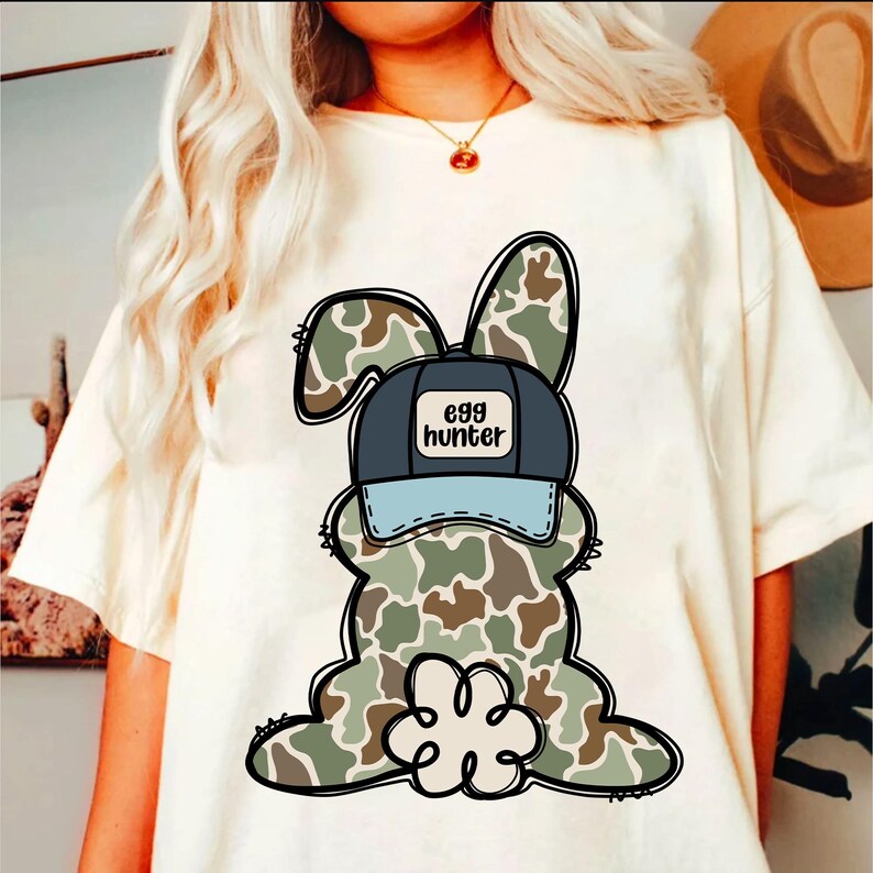 May include: White t-shirt featuring a cartoon camouflage bunny wearing a blue and white baseball cap that says "egg hunter".