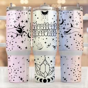 May include: Three stainless steel tumblers with a black and white celestial design. The tumblers have a gradient background with pink, yellow, and blue colors. The tumblers feature a hand holding a sun, a hand holding a moon, and a crystal. The text "Manifest That Shit" is printed on the middle tumbler.
