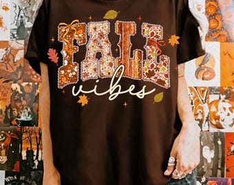 May include: A brown t-shirt with the words "FALL vibes" in a fall-themed design. The letters "FALL" are filled with floral and leaf patterns in shades of orange, brown, and cream. The word "vibes" is written in a cursive font.