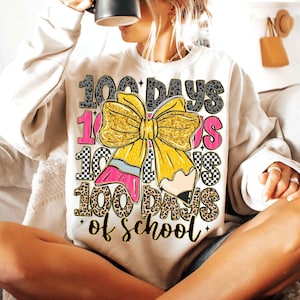 Puede incluir: Sudadera color crema con un gr&aacute;fico colorido que celebra 100 d&iacute;as de escuela. El dise&ntilde;o incluye un gran lazo amarillo con purpurina dorada, un l&aacute;piz rosa y el texto "100 Days of School" en varias fuentes y patrones.
