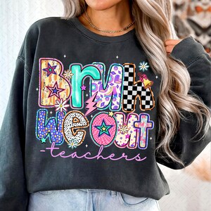 Op de afbeelding: Een donkergrijs sweatshirt met een kleurrijke grafische print die "Bring We Out - teachers" zegt. De grafiek is gemaakt van verschillende patronen en kleuren, waaronder sterren, bliksem en bloemen.