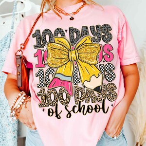 Puede incluir: Camiseta rosa con un dise&ntilde;o gr&aacute;fico que celebra los 100 d&iacute;as de escuela. El dise&ntilde;o incluye el texto "100 Days of School", un gran lazo amarillo y un l&aacute;piz, con varios patrones.