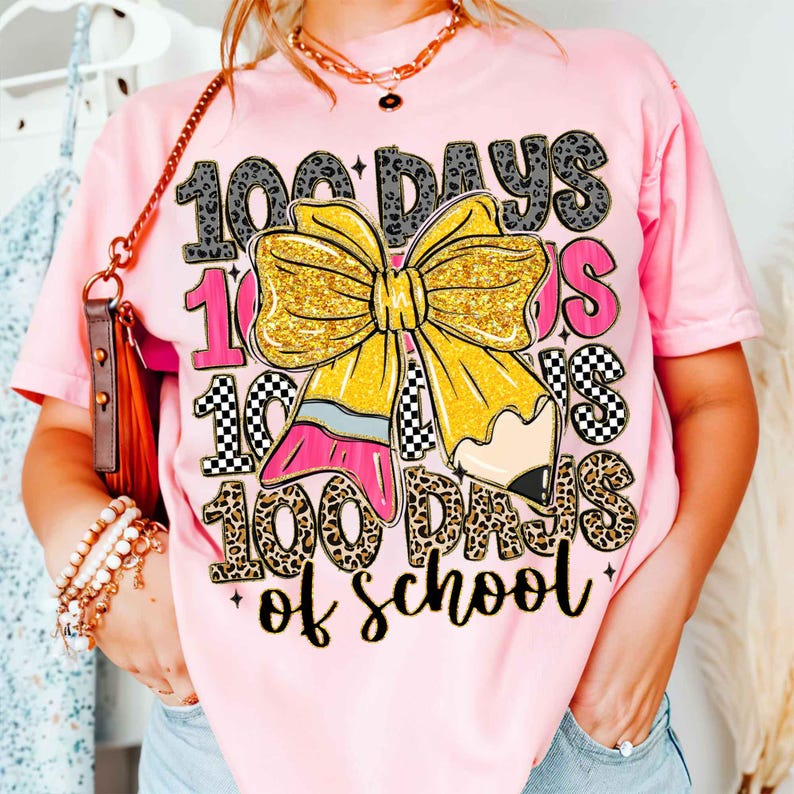 Puede incluir: Camiseta rosa con un dise&ntilde;o gr&aacute;fico que celebra los 100 d&iacute;as de escuela. El dise&ntilde;o incluye el texto "100 Days of School", un gran lazo amarillo y un l&aacute;piz, con varios patrones.