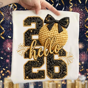 Puede incluir: Camiseta blanca con el texto bordado "hello 26". El número 26 es negro y dorado, con un lazo dorado y una esfera dorada. El diseño incluye fuegos artificiales dorados y purpurina.