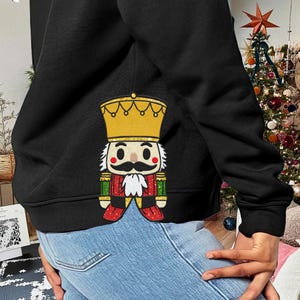 Könnte beinhalten: Schwarzes Sweatshirt mit einem farbenfrohen Nussknacker-Design auf dem Rücken. Der Nussknacker hat eine goldene Krone, einen weißen Schnurrbart und eine rot-grüne Uniform. Das Design ist glitzernd und festlich, perfekt für die Weihnachtszeit.
