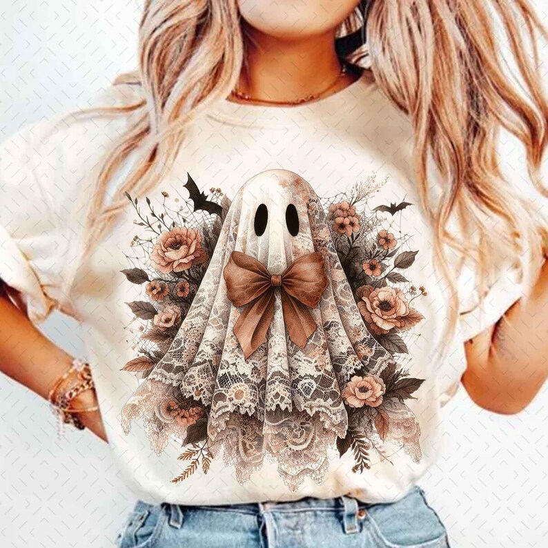以下が含まれることがあります： レースのリボンをつけた茶色と白の幽霊が描かれた白いTシャツ。幽霊は茶色の花と葉に囲まれている。