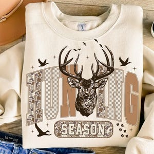 K&ouml;nnte beinhalten: Cremefarbenes Sweatshirt mit einem Hirschkopf-Motiv und den Worten "HUNTING SEASON" in braunen und camouflagefarbenen Buchstaben. Das Design umfasst fliegende V&ouml;gel und Kreuzsymbole, die eine &Auml;sthetik zum Thema Jagd schaffen.