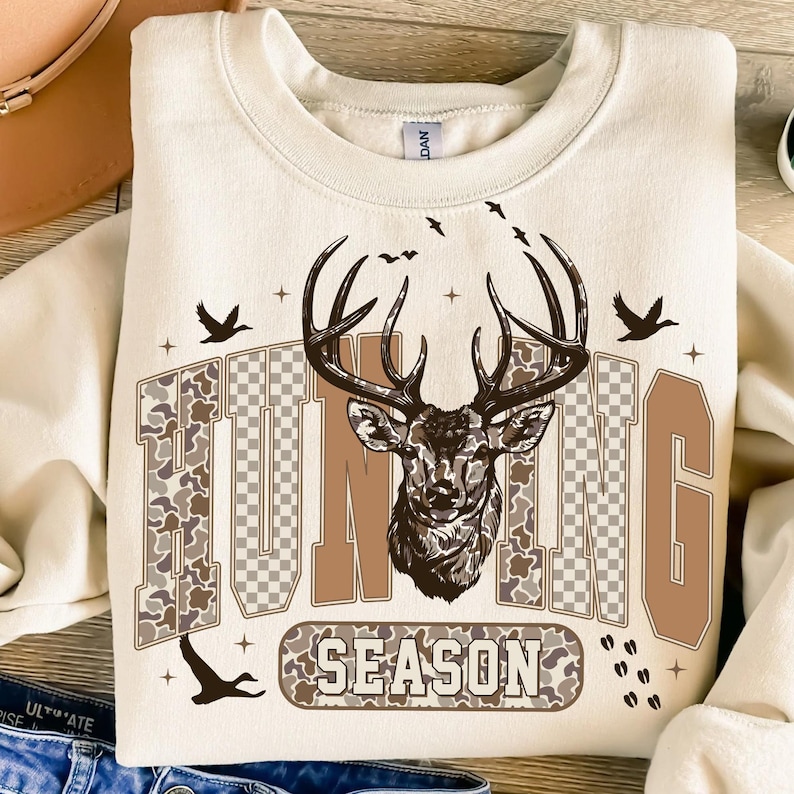 K&ouml;nnte beinhalten: Cremefarbenes Sweatshirt mit einem Hirschkopf-Motiv und den Worten "HUNTING SEASON" in braunen und camouflagefarbenen Buchstaben. Das Design umfasst fliegende V&ouml;gel und Kreuzsymbole, die eine &Auml;sthetik zum Thema Jagd schaffen.