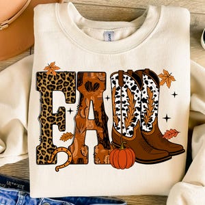May include: Cream-colored sweatshirt with the word "FALL" in a fall-themed design. The "F" is leopard print, the "A" has a cactus pattern, and the "L" is a horseshoe. Cowboy boots and a pumpkin complete the design.