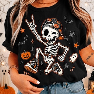 May include: A black t-shirt featuring a graphic of a skeleton wearing a pumpkin-patterned baseball cap and sneakers. The skeleton is making a rock-and-roll hand gesture, surrounded by orange stars, black bats, a jack-o'-lantern, and a small ghost.