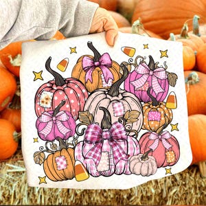 May include: A decorative pillow featuring a collection of pumpkins in shades of pink, orange, and cream. Each pumpkin has a decorative bow and a patch. Candy corn and star accents complete the fall-themed design.