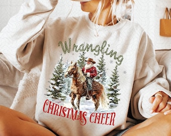 May include: A white sweatshirt with a festive design featuring a cartoon Santa Claus riding a horse through a snowy forest. The text "Wrangling Christmas Cheer" is printed on the front of the sweatshirt.