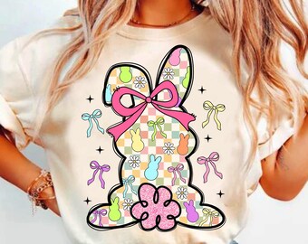 May include: A white t-shirt with a colorful graphic of a bunny made of pastel-colored Peeps candy. The bunny has a pink bow and is surrounded by small pastel-colored bows and flowers.