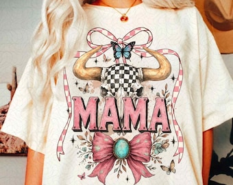 Puede incluir: Camiseta blanca con un lazo a cuadros rosa y blanco, una calavera con cuernos y la palabra "MAMA" en letras rosas. La calavera tiene un patrón a cuadros y está rodeada de mariposas y flores.