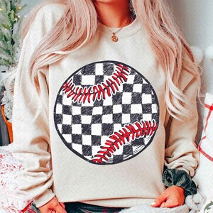 May include: A beige sweatshirt featuring a large, circular graphic of a baseball. The baseball design has a black and white checkered pattern with red stitching details. The sweatshirt has long sleeves.