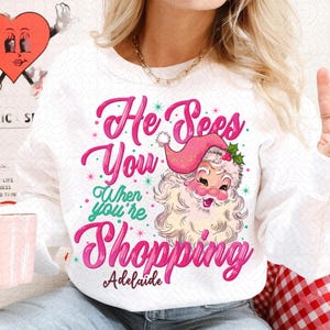 May include: White sweatshirt with a pink and green design featuring a cartoon Santa Claus and the text "He Sees You When You're Shopping Adelaide".