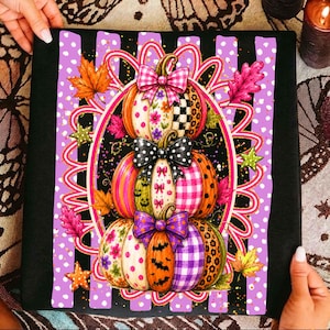 May include: A black fabric square featuring a central graphic of three stacked pumpkins decorated with various patterns including plaid, polka dots, leopard print, and floral motifs, accented by bows and colorful autumn leaves.