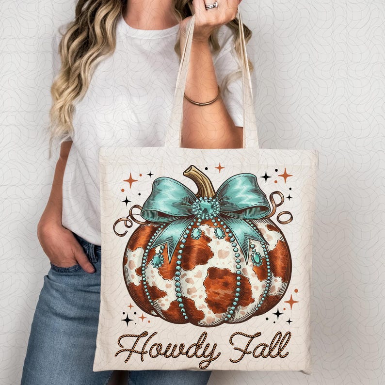 Puede incluir: Una bolsa de tela blanca con una calabaza de estampado de vaca marr&oacute;n y blanco con un lazo turquesa y pedrer&iacute;a. El texto "Howdy Fall" est&aacute; impreso en marr&oacute;n en la bolsa.