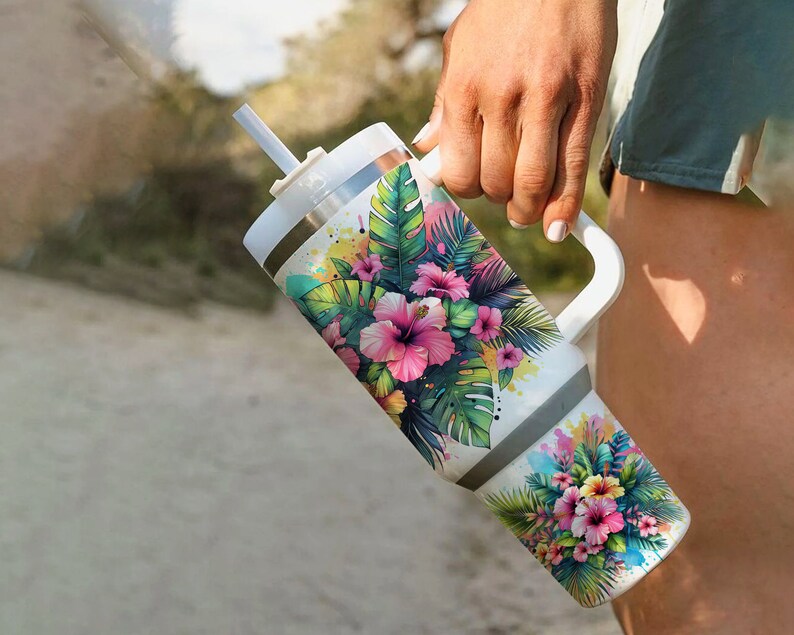 Pu&ograve; includere: Un bicchiere in acciaio inossidabile bianco con un design floreale tropicale che presenta fiori di ibisco rosa, foglie verdi e un tocco di vernice gialla. Il bicchiere ha un coperchio e un manico.