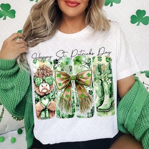 Peut inclure: T-shirt blanc avec un motif graphique vert, marron et blanc représentant une vache des Highlands de dessin animé portant des lunettes de soleil, un nœud et un trèfle. Le texte "Happy St. Patricks Day" est imprimé au-dessus du graphique.