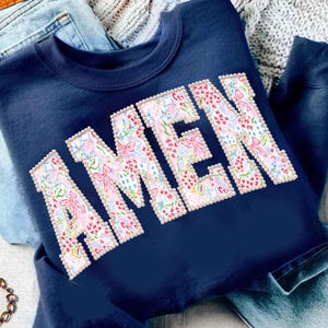 Pode incluir: Camisola azul marinho com a palavra "AMEN" em letras grandes com padrão floral, contornadas com pérolas falsas. O padrão floral inclui laços rosa, flores e um fundo azul claro. A camisola está dobrada sobre ganga.