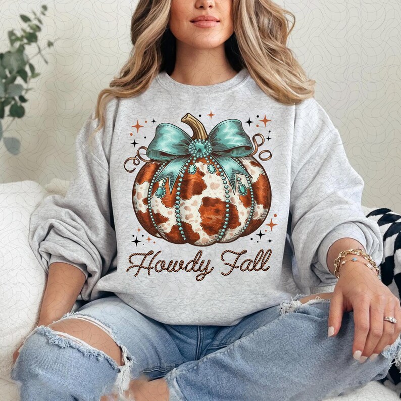 Puede incluir: Una sudadera gris con una calabaza de estampado de vaca marr&oacute;n y blanco con un lazo turquesa y pedrer&iacute;a. El texto "Howdy Fall" est&aacute; impreso debajo de la calabaza.