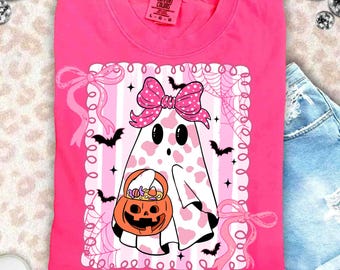 May include: A bright pink t-shirt featuring a cartoon ghost with a pink bow, holding a jack-o'-lantern candy bucket. The ghost has a pink cow-print design. The shirt has a white and pink striped background with bats and spiderwebs.