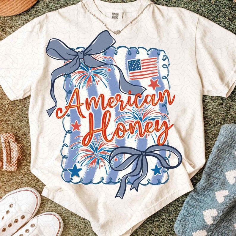 May include: Cream-coloured t-shirt with a patriotic design. The graphic features a blue and white striped rectangle with fireworks, stars, and an American flag. The words "American Honey" are written in red script, with two blue bows.