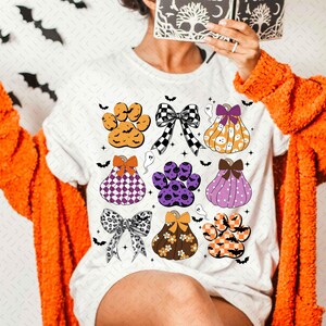 May include: White t-shirt with a Halloween-themed design featuring paw prints, pumpkins, and bows in various colors and patterns.