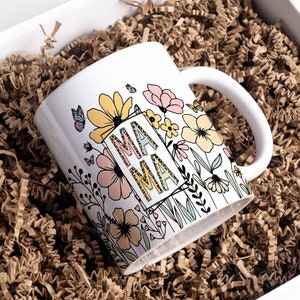 May include: White ceramic mug with a floral design and the word "MAMA" in a leopard print font. The flowers are pink, yellow, and green.