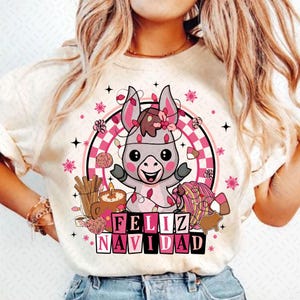 May include: A white t-shirt with a pink and gray cartoon donkey wearing Christmas lights and a flower crown. The donkey is surrounded by pink and white Christmas decorations and the text "Feliz Navidad" in pink letters.