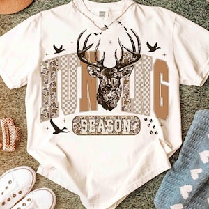 K&ouml;nnte beinhalten: Wei&szlig;es T-Shirt mit einem Hirschkopf-Motiv und den Worten "HUNTING SEASON" in braunen und Camouflage-Buchstaben. Das Design beinhaltet Silhouetten von V&ouml;geln und Hirschspuren. Eine silberne Halskette ist oben sichtbar.