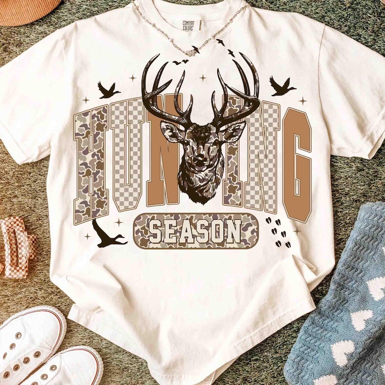 K&ouml;nnte beinhalten: Wei&szlig;es T-Shirt mit einem Hirschkopf-Motiv und den Worten "HUNTING SEASON" in braunen und Camouflage-Buchstaben. Das Design beinhaltet Silhouetten von V&ouml;geln und Hirschspuren. Eine silberne Halskette ist oben sichtbar.