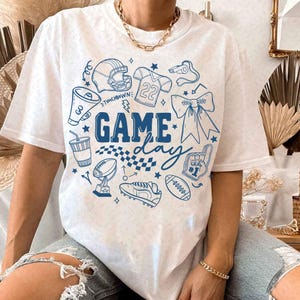 Puede incluir: Camiseta blanca con un diseño gráfico azul que presenta ilustraciones relacionadas con el fútbol americano y las palabras "Game Day". El diseño incluye un casco de fútbol americano, una camiseta, un balón y otros elementos deportivos.