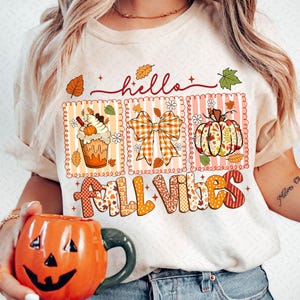 Puede incluir: Camiseta color crema con un gráfico temático de otoño. El gráfico incluye la palabra "hello" en cursiva y las palabras "Fall Vibes" en un diseño de patchwork. El gráfico también presenta un café con leche de calabaza, un lazo y una calabaza.
