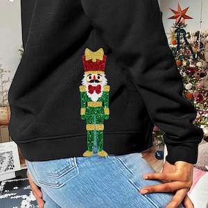 Puede incluir: Una sudadera negra con un diseño de cascanueces de purpurina verde y roja en la parte delantera. El cascanueces lleva un gorro rojo con un lazo dorado y un cinturón dorado.