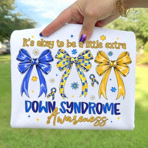 May include: White t-shirt with a design featuring three decorative bows in blue, yellow, and gold, with puzzle piece accents. The text reads "It's okay to be a little extra" and "Down Syndrome Awareness".