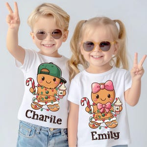 May include: Two children wearing white t-shirts with gingerbread person designs. The boy's shirt says "Charlie" and the girl's shirt says "Emily".