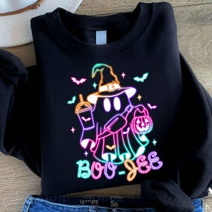 May include: Black sweatshirt featuring a neon pink, blue, and green graphic of a ghost wearing a witch hat and holding a pumpkin and a drink. The text "Boo-Jee" is written below the ghost.