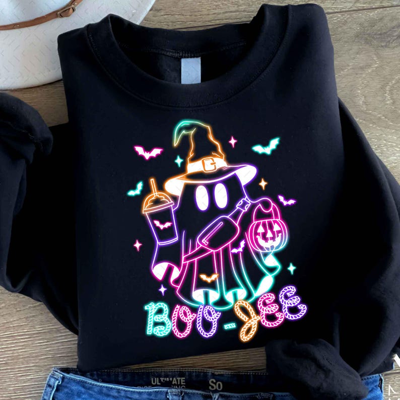 May include: Black sweatshirt featuring a neon pink, blue, and green graphic of a ghost wearing a witch hat and holding a pumpkin and a drink. The text "Boo-Jee" is written below the ghost.