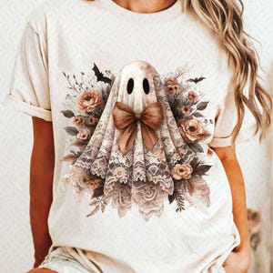 以下が含まれることがあります： レースのベールと茶色のリボンをつけた幽霊が描かれた白いTシャツ。幽霊は茶色の花と葉に囲まれている。