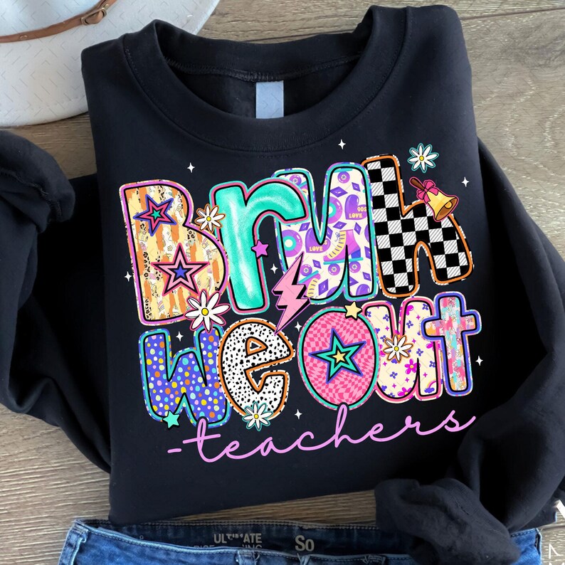 Op de afbeelding: Zwart sweatshirt met een kleurrijke afbeelding met de tekst "Bruh we out - teachers". De afbeelding is gemaakt van verschillende patronen en kleuren, waaronder sterren, bloemen en ruitjes.