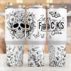 Puede incluir: Tres vasos de acero inoxidable con diferentes diseños. El primer vaso presenta una calavera de azúcar en blanco y negro con detalles florales. El segundo vaso tiene un diseño en blanco y negro con el texto "Zero Fucks Given". El tercer vaso tiene un diseño de mariposa en blanco y negro con detalles florales.