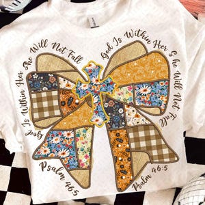 Peut inclure: T-shirt blanc avec un motif de nœud patchwork et une croix florale. Le nœud comprend divers motifs et couleurs, avec le texte "Dieu est en elle, elle ne tombera pas, Psaume 46:5."