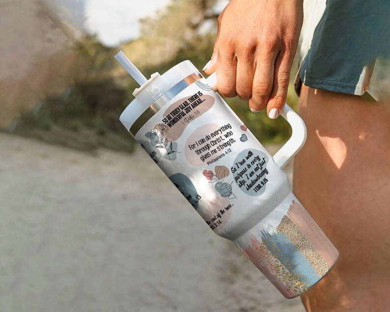 K&ouml;nnte beinhalten: Ein wei&szlig;er Becher mit einem rosafarbenen, blauen und goldenen Design. Der Becher hat einen Strohhalm und einen Griff. Er zeigt inspirierende Zitate und Bibelverse, darunter "So kann ich alles durch Christus tun, der mir Kraft gibt." und "Ich &uuml;berlebe nicht nur, ich gedeihe."