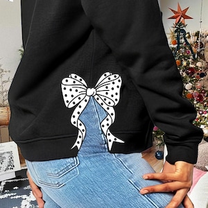 Puede incluir: Sudadera negra con un lazo blanco de lunares en la espalda. El lazo está colocado para que parezca que está atado a la cintura.