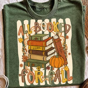 May include: A heathered olive green t-shirt featuring a graphic print of a stack of five books and an orange pumpkin. The text "ALL BOOKED FOR FALL" is printed in a multi-colored, rounded font. The design includes autumn leaves and floral accents.