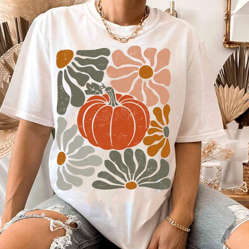 May include: White t-shirt featuring a retro-style design with a large orange pumpkin and stylized flowers in shades of orange, pink, and gray. The design has a distressed, vintage look. The shirt is a casual, everyday wear item.