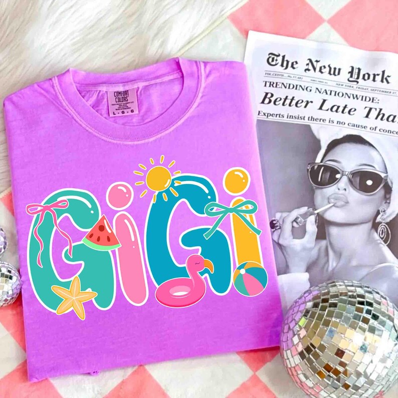 May include: A lilac Comfort Colors t-shirt with the name "GIGI" in colorful, playful lettering. The design includes a sun, watermelon slice, starfish, and a flamingo floatie. A newspaper and disco ball are in the background.