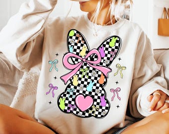May include: A white sweatshirt with a checkered bunny design. The bunny has a pink bow and is surrounded by pastel-colored Easter eggs and flowers.